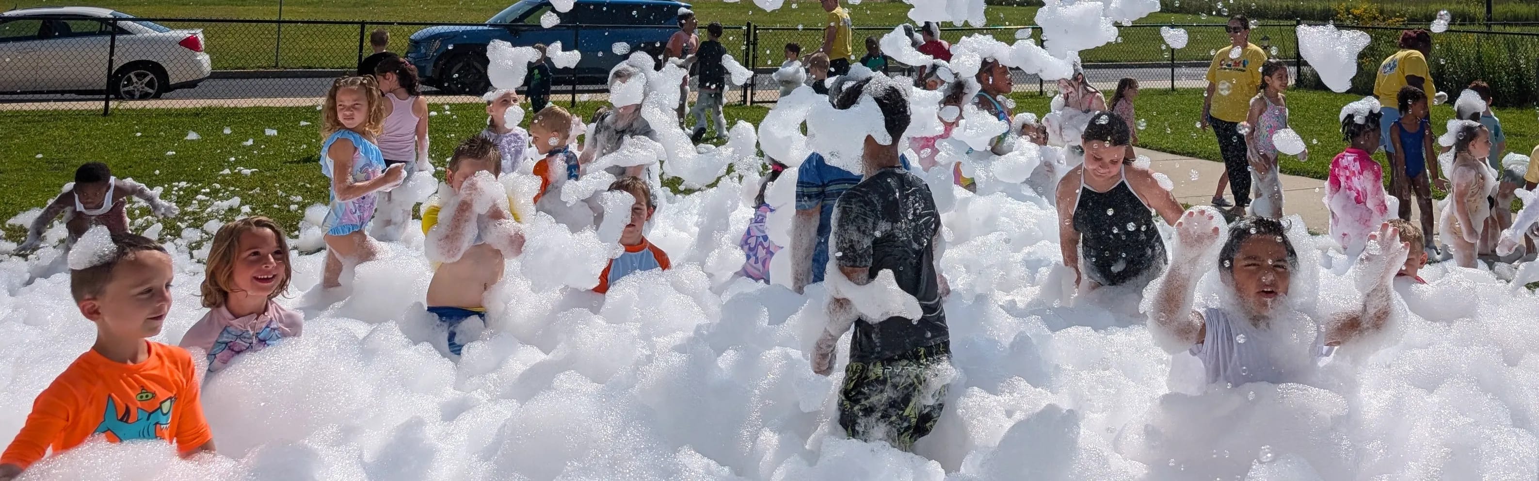 A big group of kids cheering and playing chest-deep in a wide field of foam at a sunny outdoor event