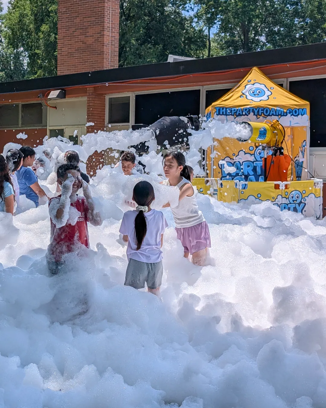 Kids playing in a large field of foam beside the Party Foam cannon and branded tent at a school event